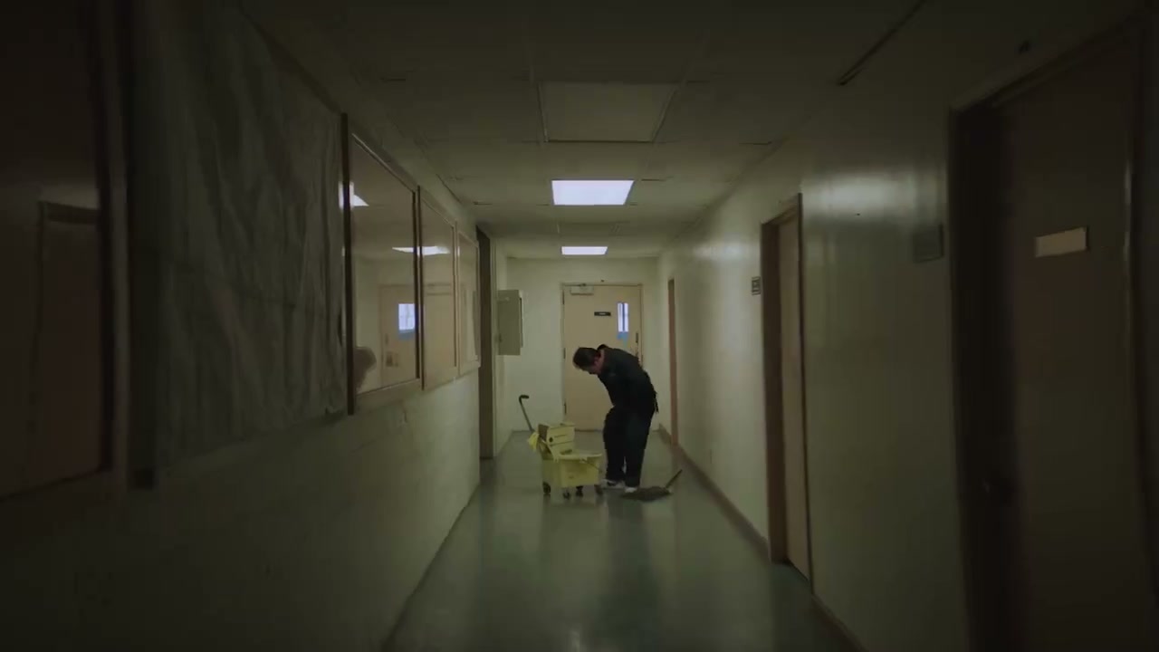 Frame from the editing reel — a dim hallway under fluorescent panels, a janitor mopping the floor